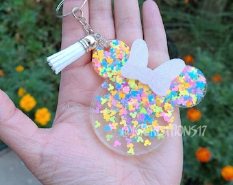 May include: A colorful, glittery resin keychain shaped like Mickey Mouse ears with a white bow. The keychain is a light pink color with a mix of pastel blue, yellow, and green glitter.