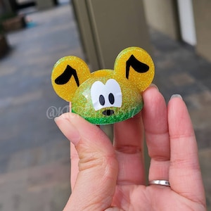 May include: A yellow and green glitter resin figurine of a cartoon dog with black ears and eyes. The figurine has a green glitter base.