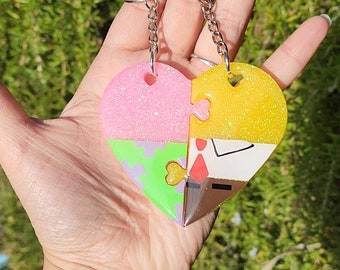 May include: A heart-shaped keychain with a puzzle piece design. The keychain is split into four sections, each with a different color and pattern: pink glitter, green with purple spots, yellow glitter, and white with a red tie. The keychain is attached to a silver chain.