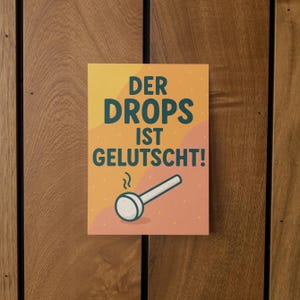 May include: A rectangular poster with the text "DER DROPS IST GELUTSCHT!" in teal letters on an orange and pink background. A white and teal pipe illustration is below the text. The poster is mounted on a wooden surface.