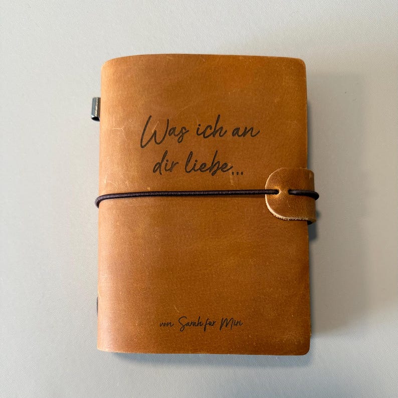 May include: A brown leather journal with a black elastic closure. The cover features the German text "Was ich an dir liebe..." and "von Sarah f&uuml;r Mia" in black script.