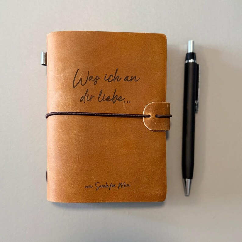 May include: A tan leather journal with the text "Was ich an dir liebe..." and "von Sarah f&uuml;r Min" printed on the cover. A black pen rests beside the journal. The journal is closed with an elastic band.