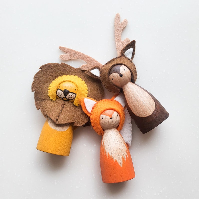 May include: Wooden peg dolls dressed as a lion, fox, and deer. The lion has a brown felt mane and yellow face. The fox is orange with white accents, and the deer has brown felt antlers. These whimsical figures are perfect for imaginative play.