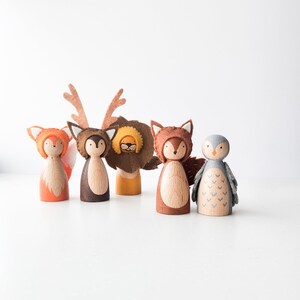 May include: Wooden peg dolls dressed as woodland animals, including a fox, deer, lion, another fox, and an owl. The dolls have felt hats and accessories in shades of orange, brown, and gray. The dolls are arranged on a white surface.