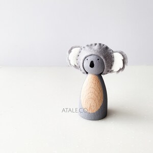 May include: A grey felt koala doll with white felt ears and a wooden body. The doll is wearing a grey felt hat with white felt ears.