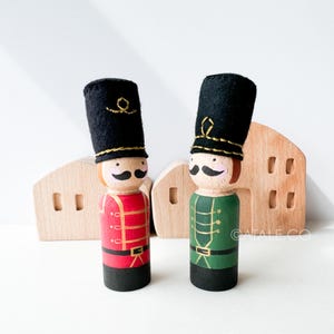 May include: Two wooden toy nutcracker soldiers with black felt hats, gold trim, and painted faces. One soldier is wearing a red uniform and the other is wearing a green uniform. The soldiers are standing in front of a wooden toy house.
