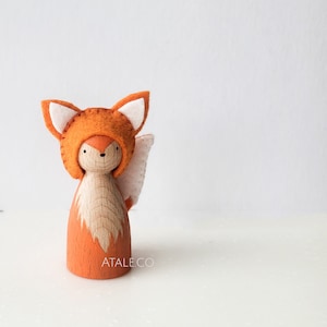 May include: A wooden peg doll dressed as a fox. The doll features a carved wooden body, an orange painted lower section, and a felt fox hood with white inner ears. A white and pink felt tail is attached to the back. The text "ATALE.CO" is printed on the doll.