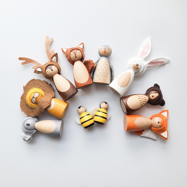 May include: A collection of wooden peg dolls, each uniquely designed as woodland creatures. The set includes a deer, fox, owl, rabbit, lion, bee, and bear, with felt accents and painted details. The dolls are arranged on a white surface.