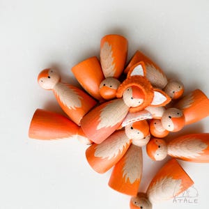 May include: A collection of orange wooden fox figurines with white and tan accents. Some figurines have fox tail designs, while others have fox faces. One figurine wears a felt fox hat. The figurines are arranged on a white surface.
