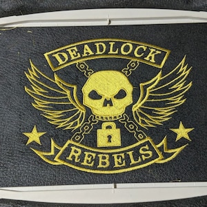 Puede incluir: Diseño bordado en tela negra con una calavera con alas, cadenas y un candado. El texto "DEADLOCK REBELS" está en una pancarta debajo de la calavera.