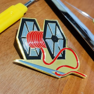 Puede incluir: Un pin de metal dorado con dos cazas TIE de Star Wars, con un carrete de hilo rojo entre ellos. Una aguja dorada con hilo rojo está unida al carrete.