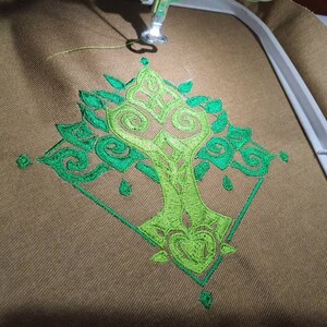 Op de afbeelding: Een geborduurd ontwerp van een gestileerde boom in groen, met ingewikkelde details, op een bruine stoffen achtergrond.