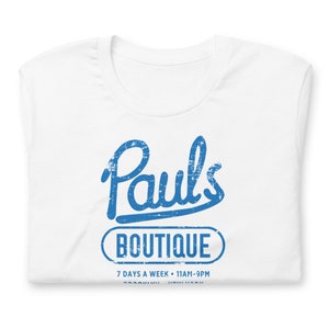 Könnte beinhalten: Weißes T-Shirt mit einem blauen, verwaschenen Aufdruck. Der Aufdruck lautet "Pauls Boutique" über einem Oval mit "Boutique" darin. Darunter steht "7 DAYS A WEEK • 11AM-9PM" und "BROOKLYN • NEW YORK".