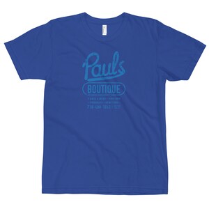 Könnte beinhalten: Ein königsblaues T-Shirt mit einer abgenutzten blauen Grafik von "Paul's Boutique" mit dem Text "7 days a week - 11AM-8PM - Brooklyn - New York - 718-498-1043" darunter.
