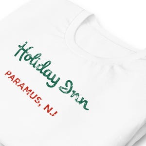 以下が含まれることがあります： 白のTシャツ。緑の筆記体で「Holiday Inn」、赤のブロック体で「PARAMUS, NJ」と書かれています。柔らかく快適な素材で作られています。