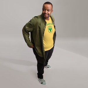 May include: A man wearing a yellow t-shirt with a green logo that says "Easties" and a green and white graphic. He is also wearing a green jacket and black pants.
