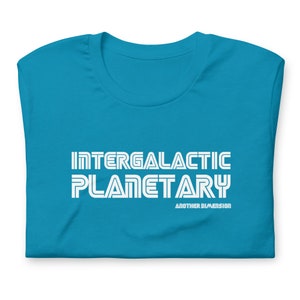 May include: A folded turquoise t-shirt with the words "INTERGALACTIC PLANETARY" in white, bold, retro-style font. Below the main text is the phrase "ANOTHER DIMENSION" in a smaller font.
