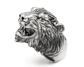 May include: A silver ring with a detailed bear head design. The bear's mouth is open, showing its teeth. The ring is made of metal and has a textured finish.