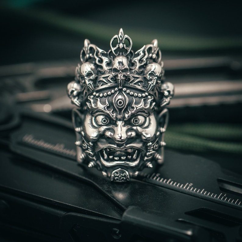 K&ouml;nnte beinhalten: Ein silberner Ring mit einem detaillierten Design eines D&auml;monengesichts mit einer Krone aus Sch&auml;deln. Der Ring liegt auf einer schwarzen Oberfl&auml;che.