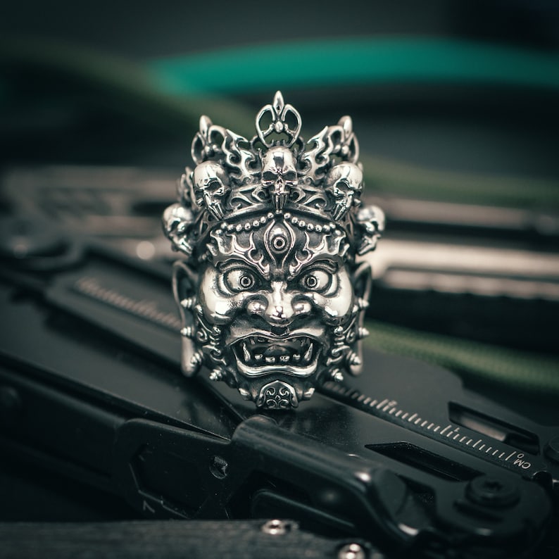 K&ouml;nnte beinhalten: Ein silberner Ring mit einem detaillierten Design eines D&auml;monengesichts mit einer Krone und Sch&auml;deln. Der Ring liegt auf einer schwarzen Oberfl&auml;che.
