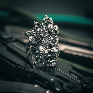 K&ouml;nnte beinhalten: Ein silberner Ring mit einem detaillierten Design einer mythischen Kreatur mit mehreren Augen und einer Krone aus Sch&auml;deln. Der Ring liegt auf einer schwarzen Oberfl&auml;che.