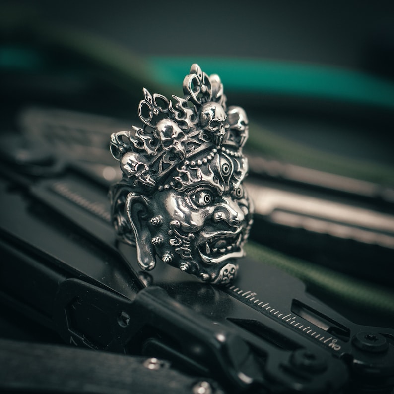 K&ouml;nnte beinhalten: Ein silberner Ring mit einem detaillierten Design einer mythischen Kreatur mit mehreren Augen und einer Krone aus Sch&auml;deln. Der Ring liegt auf einer schwarzen Oberfl&auml;che.