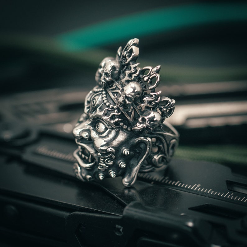 K&ouml;nnte beinhalten: Ein silberner Ring mit einem detaillierten Design eines D&auml;monengesichts mit einer Krone aus Sch&auml;deln. Der Ring befindet sich auf einem schwarzen Hintergrund.