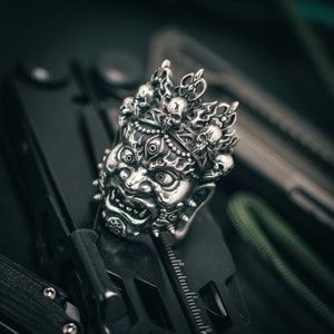 K&ouml;nnte beinhalten: Ein silberner Anh&auml;nger mit einem detaillierten, verzierten Design einer mythischen Kreatur mit einer Krone aus Sch&auml;deln. Der Anh&auml;nger ist aufwendig gearbeitet mit einem grimmigen Ausdruck und mehreren Augen.
