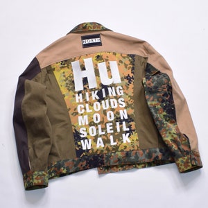 May include: A tan, olive green, and camouflage-patterned jacket with the word "NORTH" on a patch. The back of the jacket features the word "Hu" above the words "HIKING CLOUDS MOON SOLEIL WALK" in white.
