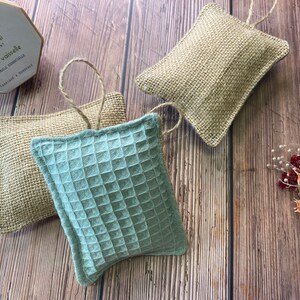 May include: Three handmade dishcloths, two are made of natural burlap and one is a light blue waffle weave fabric. The dishcloths are hanging from a string and are on a wooden surface.