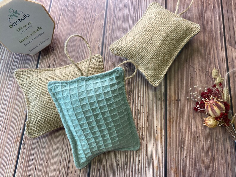 May include: Three handmade dishcloths, two are made of natural burlap and one is a light blue waffle weave fabric. The dishcloths are hanging from a string and are on a wooden surface.