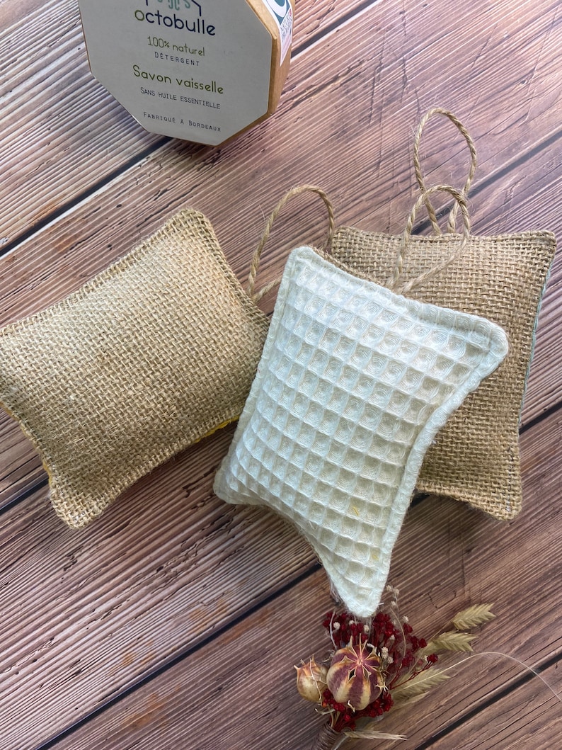 May include: Three dishcloths, two are made of burlap and one is white with a waffle texture. The dishcloths are on a wooden surface. The dishcloths are hanging from a string. A small yellow box with the text "Octobulle 100% naturel Detergent Savon vaisselle Sans huile essentielle Fabriqu&eacute; &agrave; Bordeaux" is in the upper left corner of the image.