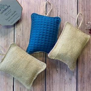 May include: Three dishcloths, two are beige and one is blue. The dishcloths are made of natural materials and have a textured surface. They are hanging from a string.