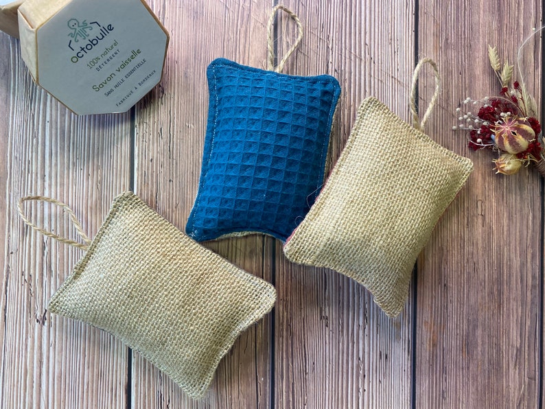 May include: Three dishcloths, two are beige and one is blue. The dishcloths are made of natural materials and have a textured surface. They are hanging from a string.