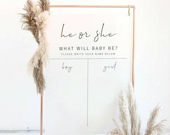 May include: A white sign with a copper frame and pampas grass on either side. The sign says "he or she? What will baby be? Please write your name below." There are two blank spaces below the text, one labeled "boy" and the other labeled "girl".