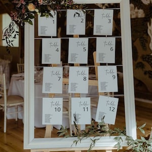 Puede incluir: Un cuadro blanco enmarcado con un plano de asientos con caligrafía negra para una recepción de boda. El cuadro se muestra en un caballete de madera y está decorado con vegetación. Los números de mesa son del 1 al 12.