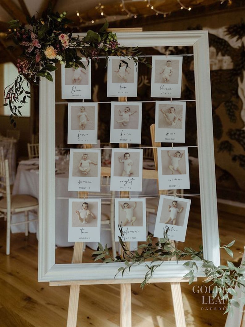 May include: A white picture frame with a white mat on a wooden easel. The frame holds a grid of photos of a baby at different ages, with each photo labeled with the number of months old the baby is. The photos are hung with clothespins and there is greenery around the frame.