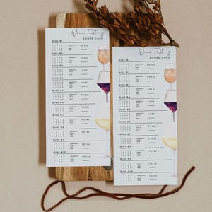May include: Two white wine tasting score cards with watercolor illustrations of wine glasses, displayed on a wooden board. The cards have the text "Wine Tasting Score Card" at the top and space to rate ten wines.