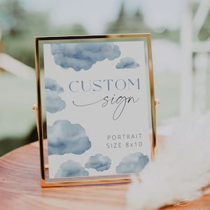 May include: A gold-framed sign with a watercolor background of blue clouds. The sign says "CUSTOM sign" and "PORTRAIT SIZE 8x10". The sign is sitting on a wooden table with a white feather plume in the foreground.