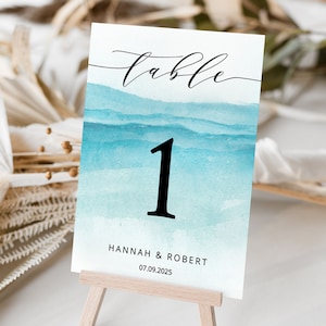 May include: Table number one with a watercolour background of blue and teal waves. The text "table" is written in cursive above the number. The names "Hannah & Robert" and the date "07.09.2025" are printed below the number.