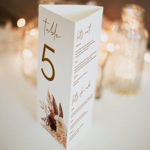 May include: Table number five with a menu for a wedding reception. The menu includes starters, main course, dessert, and drinks. The table number is in gold lettering and the menu is written in a brown font. The background is a white triangle with a floral design.