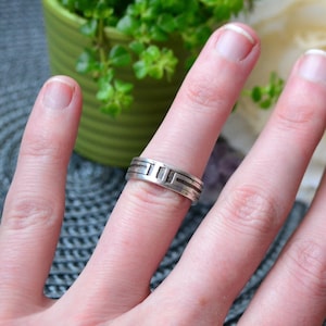 Puede incluir: Un anillo de banda de plata con un diseño geométrico. El anillo está en un dedo.