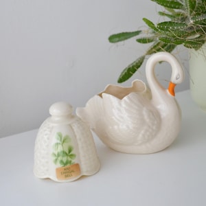 Puede incluir: Una maceta con forma de cisne de cerámica color crema con pico naranja y una campana a juego con un diseño de planta verde y el texto "4051 ERRIGAL BELL". Una planta verde en una maceta verde claro está en el fondo.