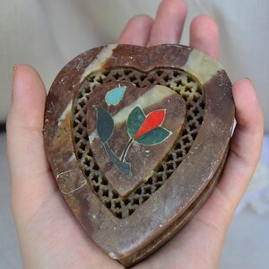 Puede incluir: Una caja de piedra en forma de coraz&oacute;n con un dise&ntilde;o floral tallado. La caja est&aacute; hecha de piedra marr&oacute;n y blanca con incrustaciones de turquesa, rojo y verde.