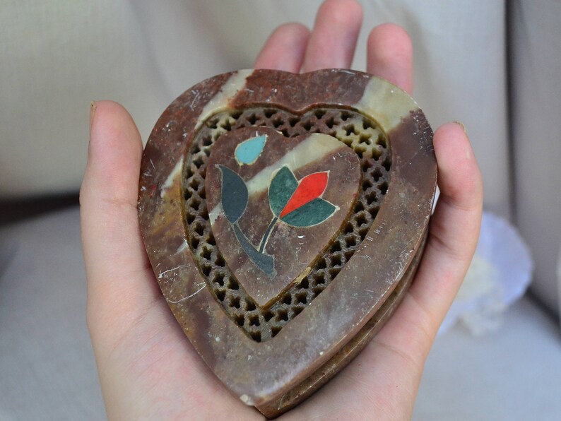 Puede incluir: Una caja de piedra en forma de coraz&oacute;n con un dise&ntilde;o floral tallado. La caja est&aacute; hecha de piedra marr&oacute;n y blanca con incrustaciones de turquesa, rojo y verde.
