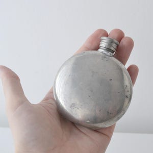 Peut inclure: Une flasque ronde en métal argenté avec une surface lisse légèrement patinée et un petit bouchon à vis. Le récipient présente un corps circulaire et un col court et étroit, affichant un éclat métallique avec quelques légères imperfections.