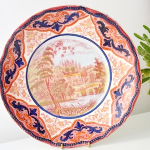 May include: Ceramic decorative plate with a scalloped edge. The plate features a central landscape scene in pink, orange, and green, surrounded by ornate blue and orange floral patterns. The rim is a deep red.