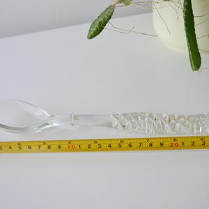 Pode incluir: Uma colher de vidro transparente com um cabo decorativo e texturizado. A colher tem aproximadamente 23 cm de comprimento. Uma fita métrica amarela é colocada ao lado da colher para escala. A colher está em uma superfície branca.