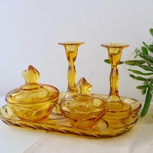 Könnte beinhalten: Ein Vintage-Schminkset aus bernsteinfarbenem Glas, bestehend aus einem Tablett, zwei Deckelgläsern und zwei Kerzenhaltern. Das Set hat ein dekoratives, strukturiertes Design und wird auf einer weißen Oberfläche präsentiert. Das Glas hat einen warmen, goldenen Farbton.