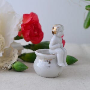 May include: A small, white porcelain figurine of a seated cherub with gold accents on its hair. The cherub sits atop a small, white pot with gold lettering. The background includes red flowers, green buds, and white lace.